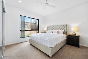 A bedroom with a large bed, a nightstand, a lamp, a ceiling fan, and a window with a view of the outside. - The Pinnacle | Luxury 4BR Home | Alfresco Living (Burpengary East)