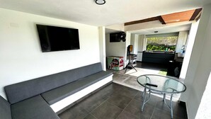 Una sala de estar moderna con un televisor de pantalla plana montado en la pared, un sofá gris oscuro, una mesa de vidrio y una cocina visible a través de un vano abierto.