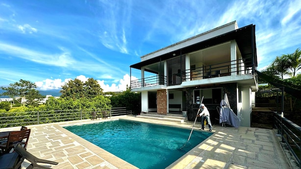 Una casa moderna de dos pisos con piscina, mobiliario exterior y una persona limpiando la piscina.