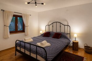 A neatly made bed with a patterned bedspread, two pillows, and rolled towels on a metal frame bed in a room with a window, a lamp, and a small wooden stool. - Two holiday homes on one estate in the heart of Podravina (Đurđevac)