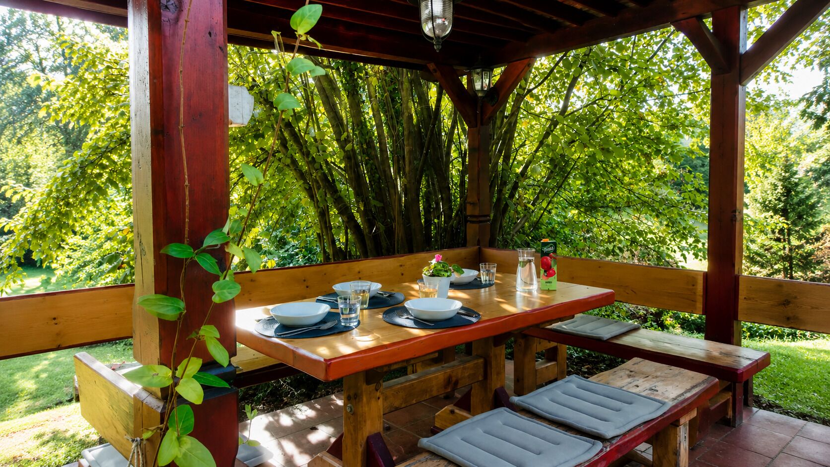 Une table en bois, dressée pour deux personnes, avec des assiettes, des bols, des couverts et des verres, entourée de bancs et d’une structure en bois offrant une vue sur une végétation luxuriante.