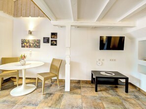 Une pièce avec un plafond blanc, un téléviseur fixé au mur, une table ronde avec un vase, des chaises en osier et une petite table noire.
