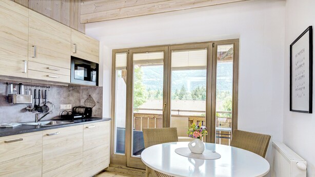 A modern kitchen with wooden cabinets, a built-in microwave, and a dining area with wicker chairs and a white table. - Edelweiss 2 by Interhome (Kaprun)