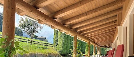 A covered outdoor seating area with wooden beams and red chairs, overlooking a gravel path and greenery.