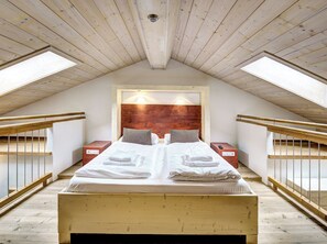 Ein Schlafzimmer mit schrägen Wänden, Holzbalken, einem großen Bett mit weißer Bettwäsche, zwei Nachttischen und einem Holz-Kopfteil.