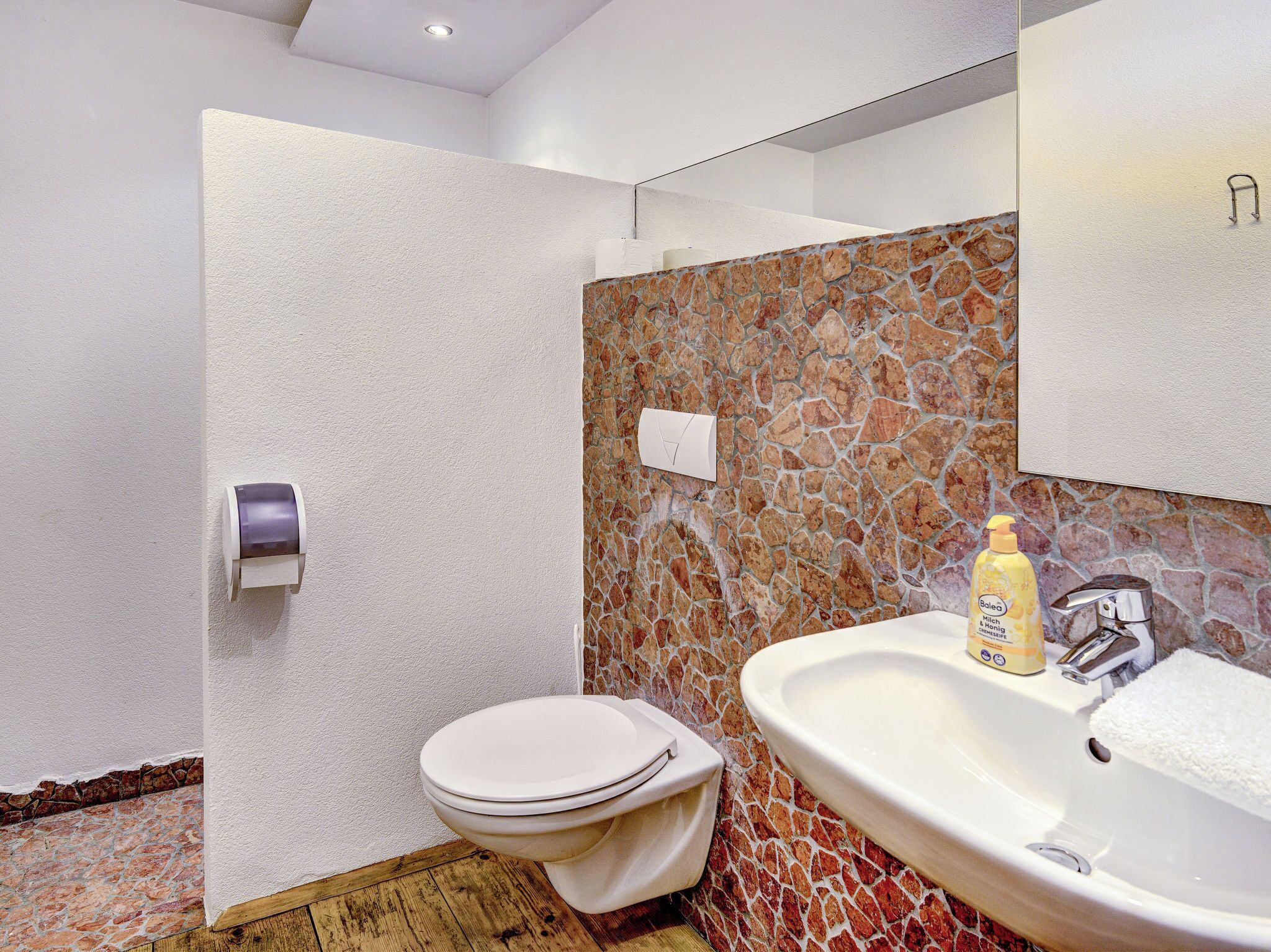 Une salle de bain moderne avec un mur en pierre, des toilettes blanches et un lavabo surmonté d’un miroir.