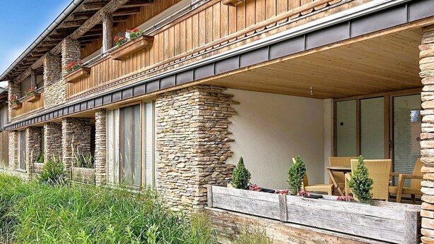 Un edificio de dos pisos con fachada de piedra y madera, balcones con jardineras de flores y una zona de estar al aire libre techada.