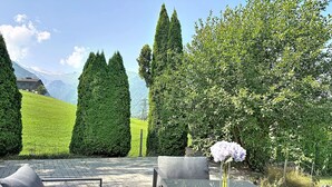 Ein Tisch mit Glasplatte, eine Vase mit Blumen, eine Couch und ein Blick auf eine gebirgige Landschaft mit ordentlich gestutzten Hecken.