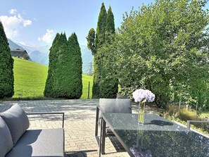 Ein Tisch mit Glasplatte, eine Vase mit Blumen, eine Couch und ein Blick auf eine gebirgige Landschaft mit ordentlich gestutzten Hecken.