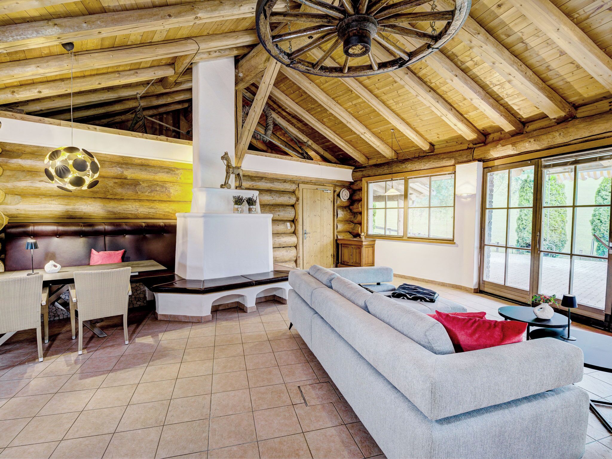 Interior de una cabaña de troncos con chimenea de piedra, un sofá, una mesa de comedor y ventanas grandes.