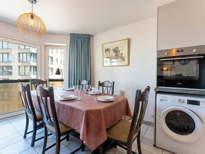 Un coin repas avec une table dressée pour quatre personnes, des chaises, un lave-linge et une fenêtre donnant sur des immeubles.