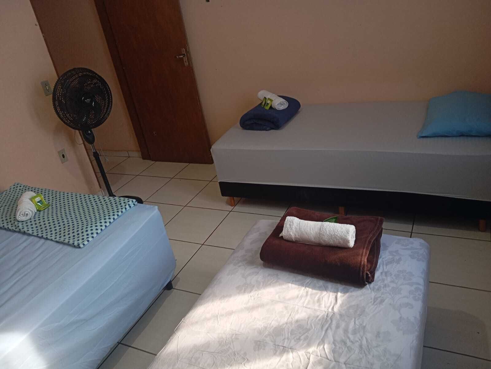 Quarto com duas camas, uma com colcha estampada e outra lisa. Há um ventilador perto de uma porta, e uma toalha está no chão.