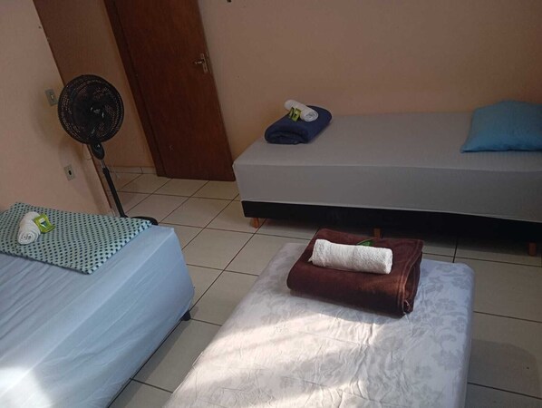 A room with two beds, one with a patterned bedspread and the other with a plain one. There is a fan near a door, and a towel is placed on the floor.