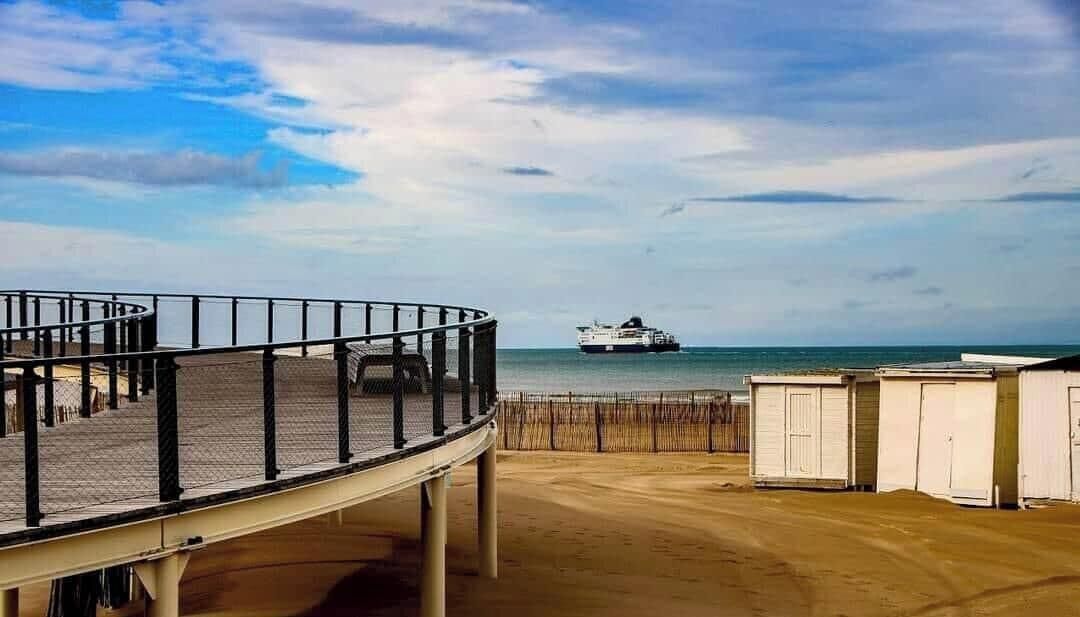 A beach with a pier, a ship in the distance, and beach huts.