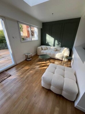 Ein modernes Wohnzimmer mit einer Couch, einem Hocker und einem Couchtisch, großen Fenstern und Blick ins Freie.