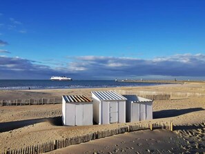 Een strand met een rij witte, verplaatsbare constructies, een schip in de verte en een houten hek.