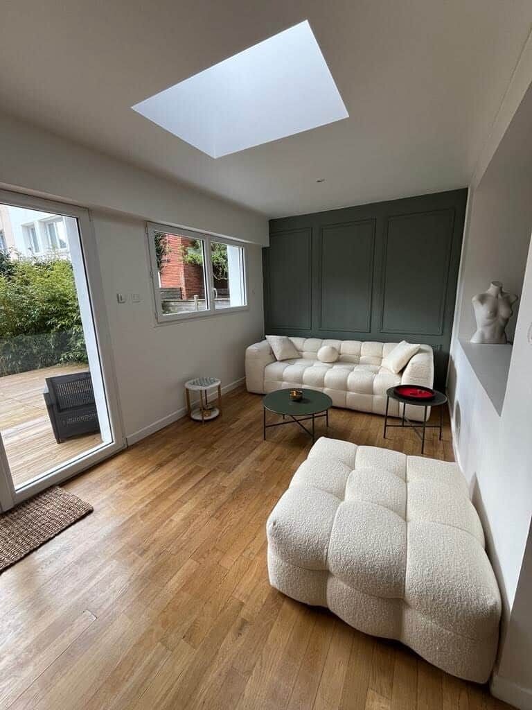 Un salon moderne avec un canapé, un pouf et une table basse, une lucarne et une vue dégagée grâce à de grandes portes vitrées.