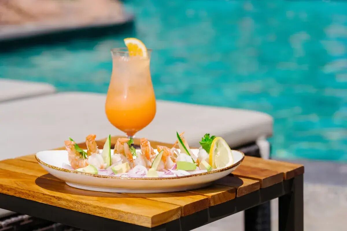 A plate of shrimp cocktail with a glass of orange drink on a wooden table by a pool.