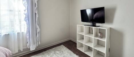 A room with a television on a white stand, a white furry rug, and sheer curtains.