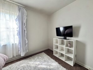 A room with a television on a white stand, a white furry rug, and sheer curtains. - Lux Modern 3BR • King Bed • SmartTV • MassageChair • Pet Friendly • Near Ft Hood (Killeen)