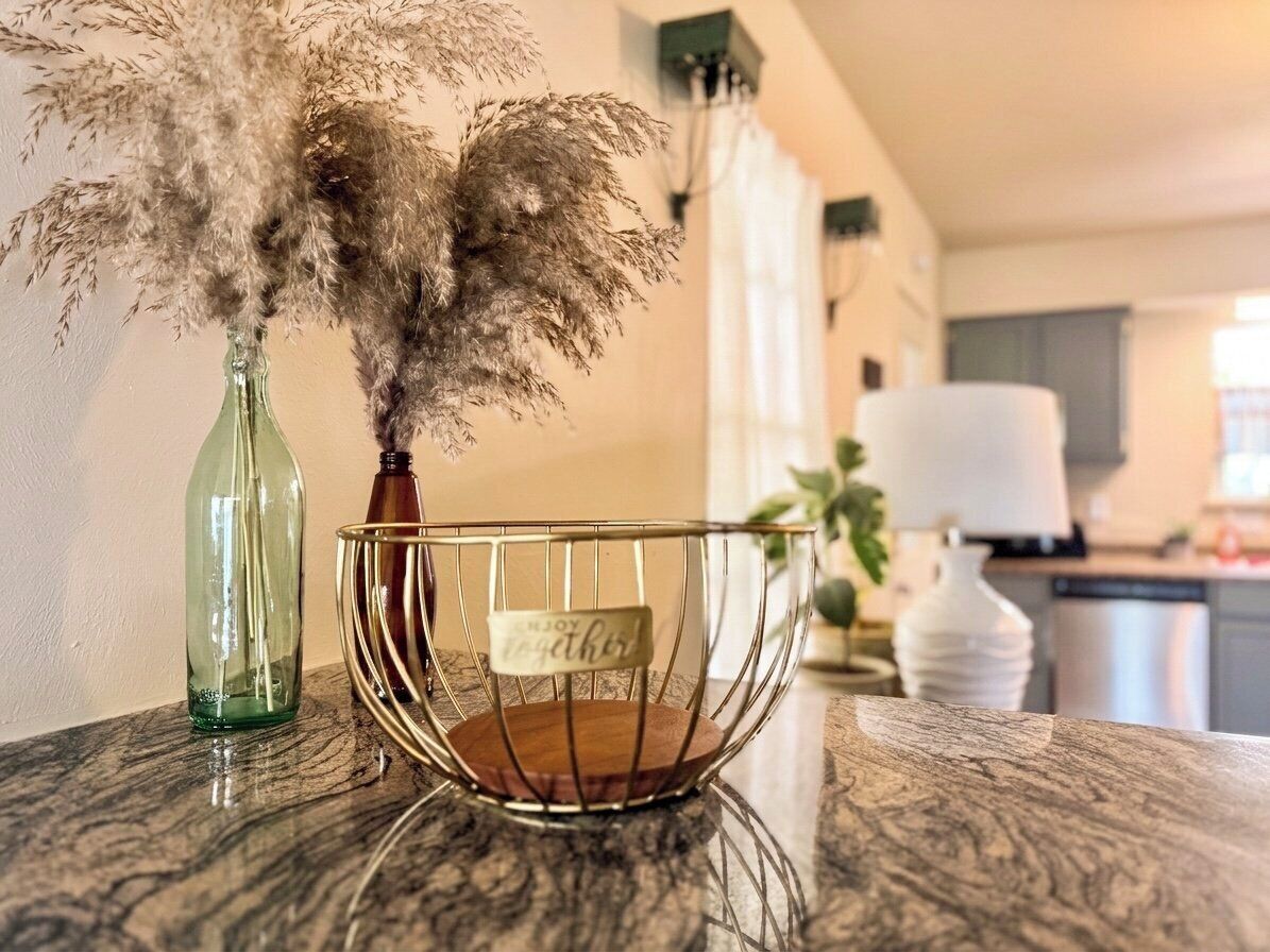 A kitchen counter with a green glass bottle, a metal bowl with a wooden base, and dried pampas grass.