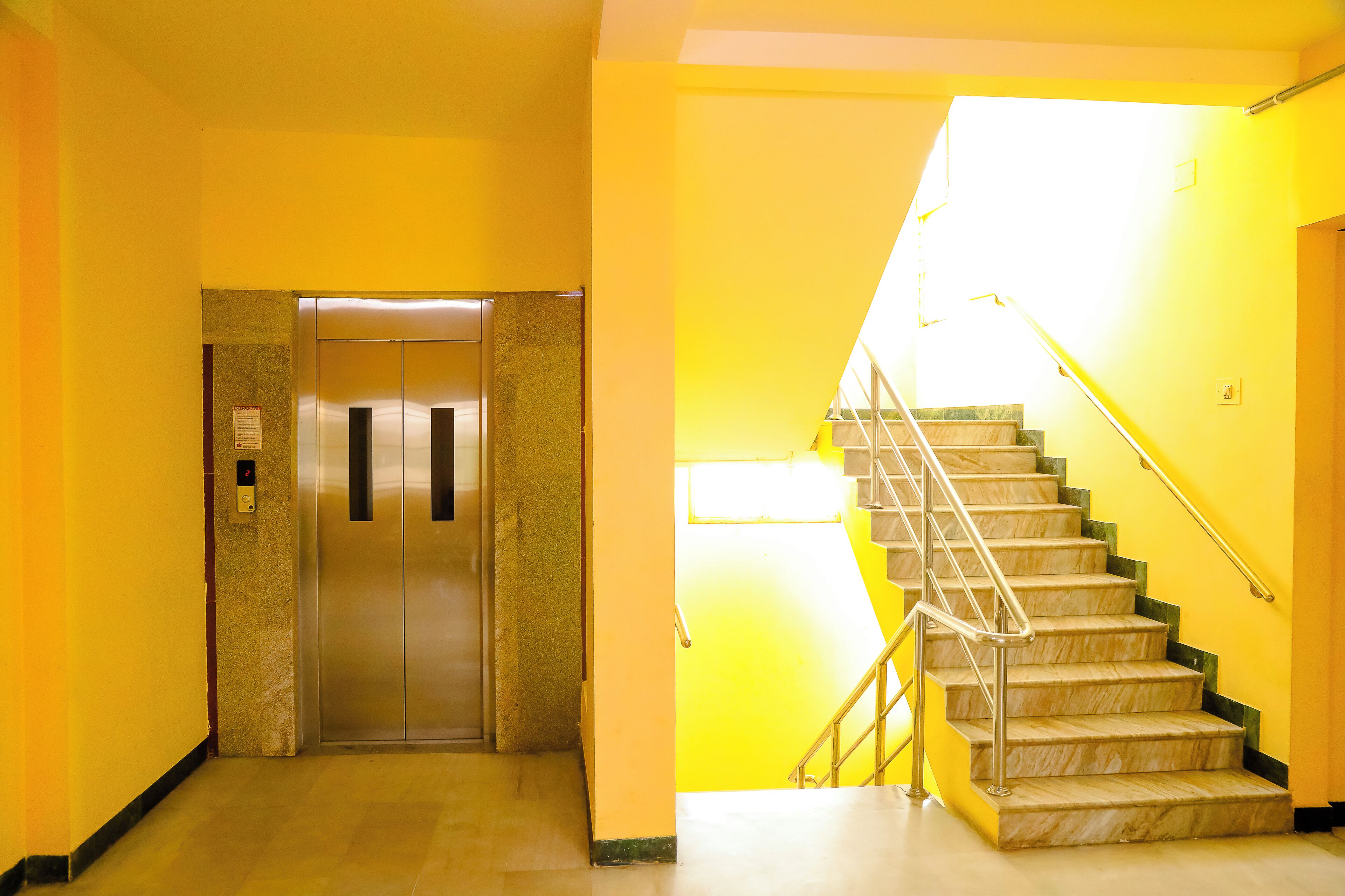 Un passadís interior amb un ascensor metàl·lic i una escala de marbre amb baranes de metall.