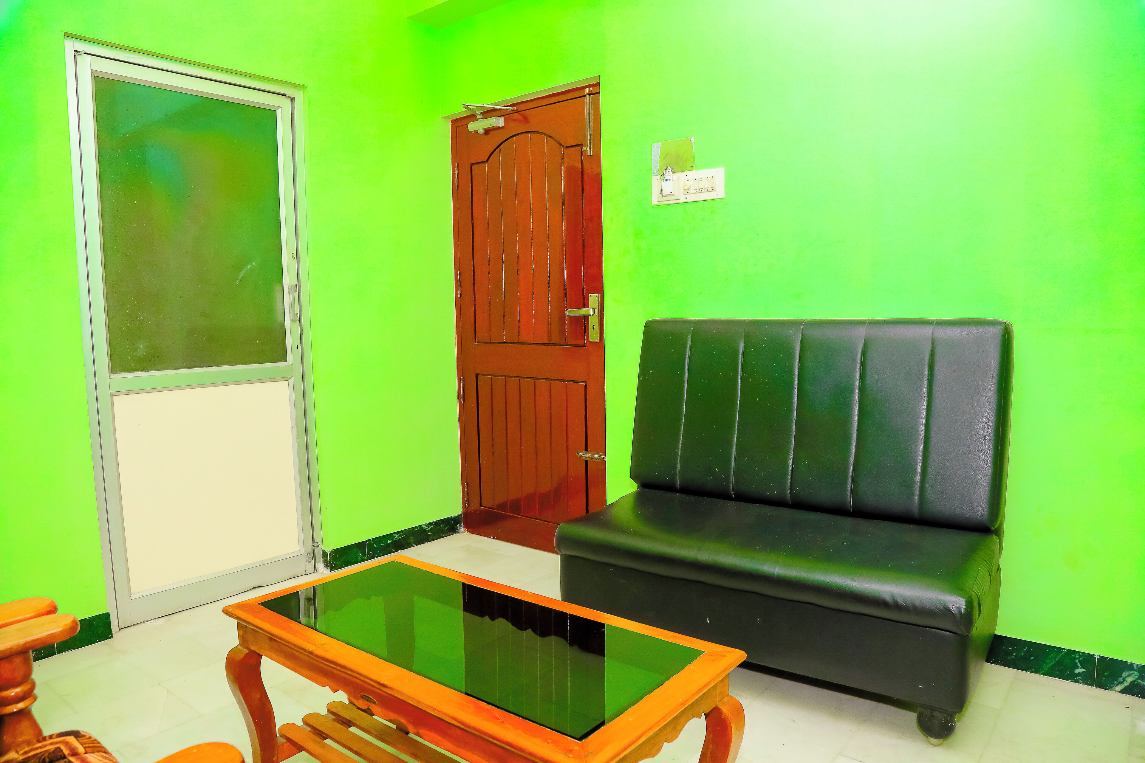 A room with a green wall, a wooden door, a black leather sofa, and a glass-top coffee table.