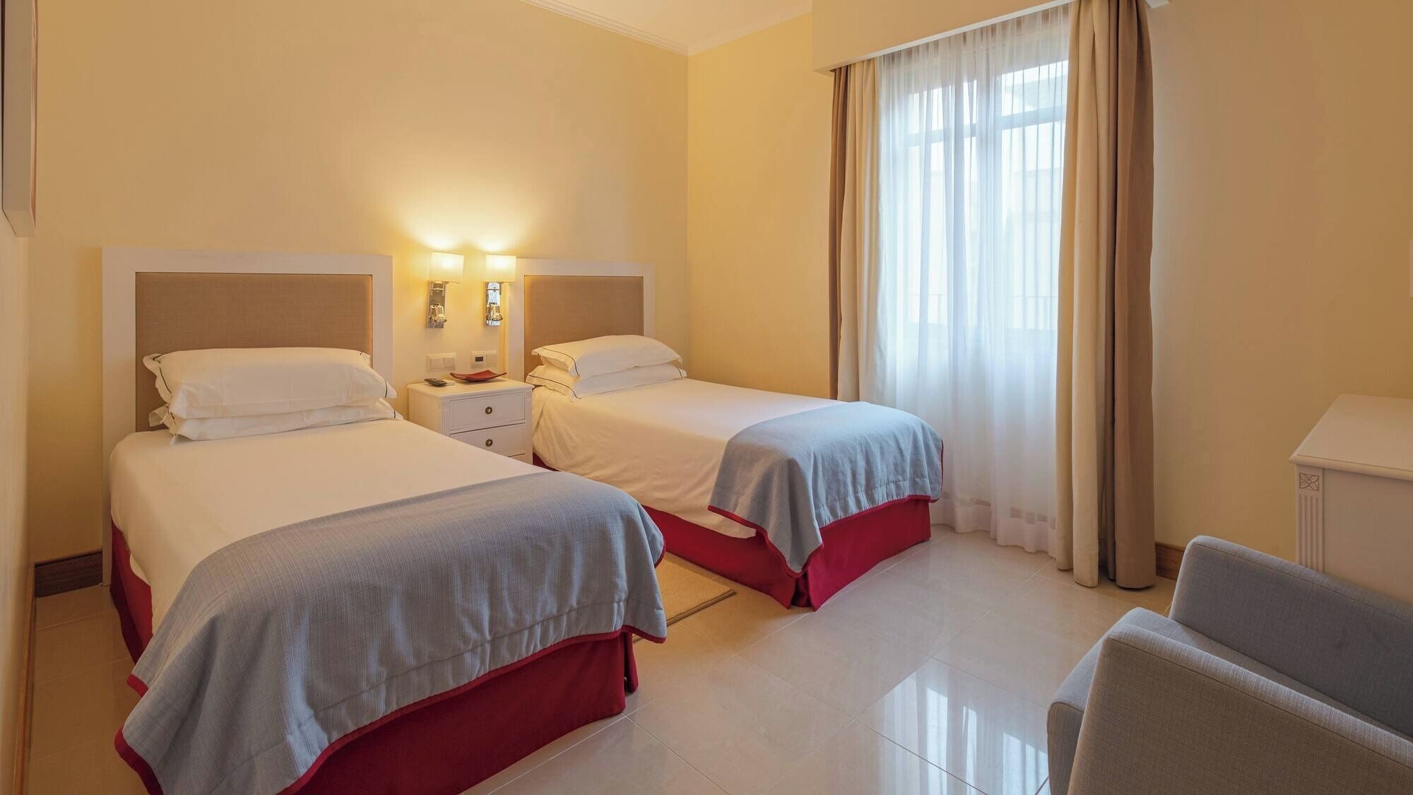 A hotel room with two beds, a nightstand, a chair, and a large window with curtains.