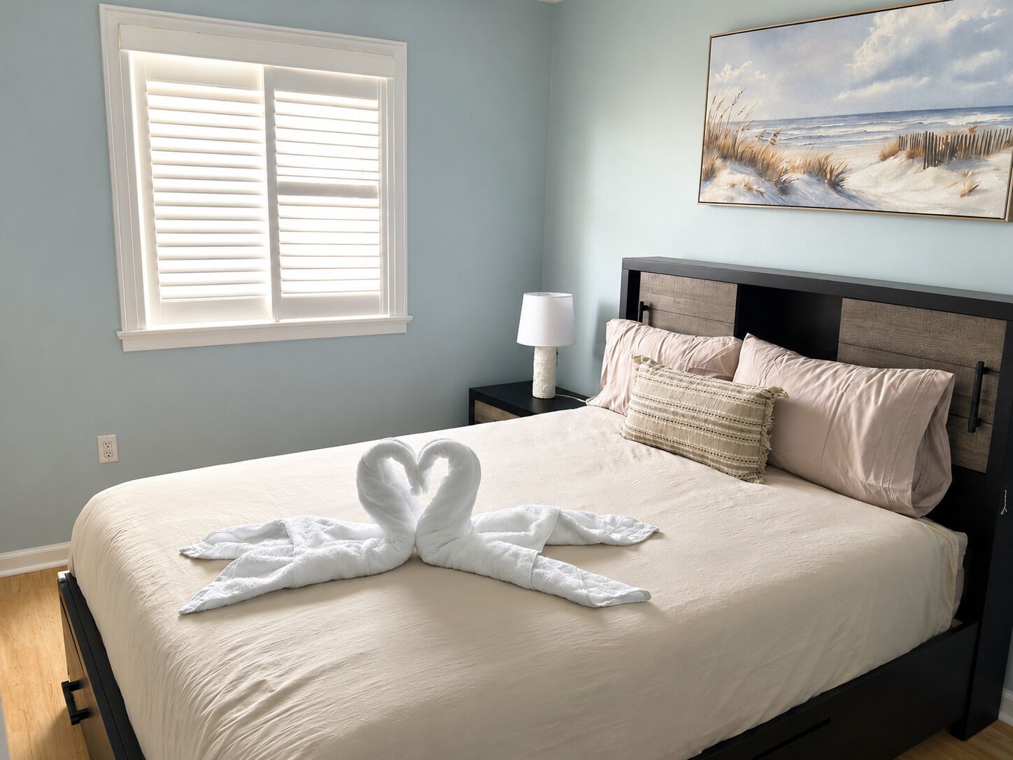 A bedroom with a bed, a nightstand, a lamp, a painting, and a window with blinds.