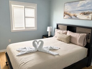 Un dormitorio con cama, mesita de noche, lámpara, un cuadro y ventana con persianas.