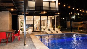 A modern outdoor pool area with a two-story building, a dining area with red chairs, and a lounge with white chairs.