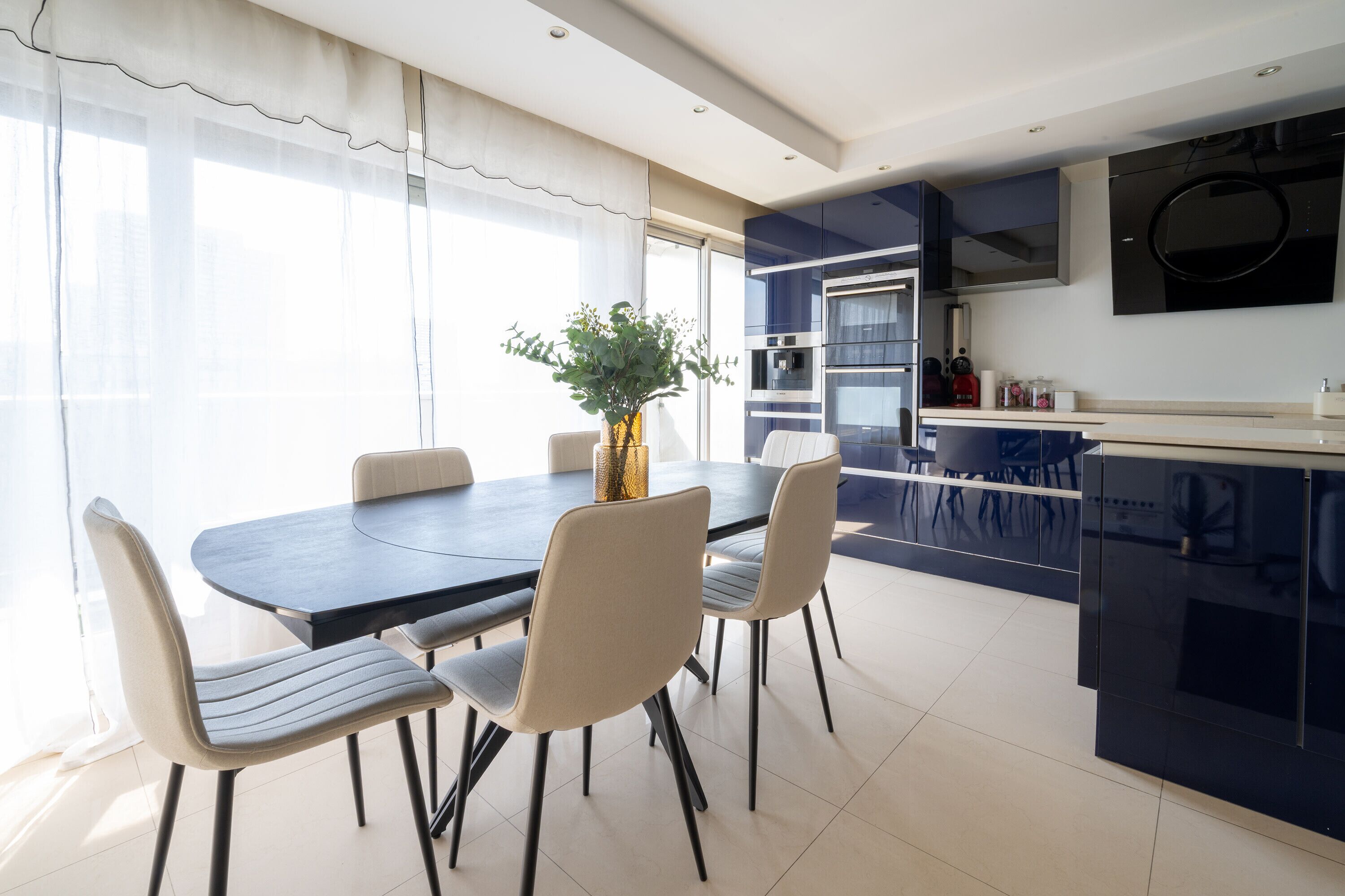 Een moderne eetruimte met een ronde tafel en vier stoelen, een potplant, en een keuken met blauwe kasten en een inbouwoven.