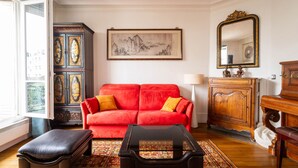 Una sala de estar con un sofá rojo, un reposapiés negro, un armario de madera, un cuadro enmarcado y un espejo.
