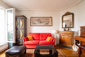 A living room with a red sofa, a black ottoman, a wooden cabinet, a framed painting, and a mirror. - Le Charonne Haussmann - Welkeys (Paris)