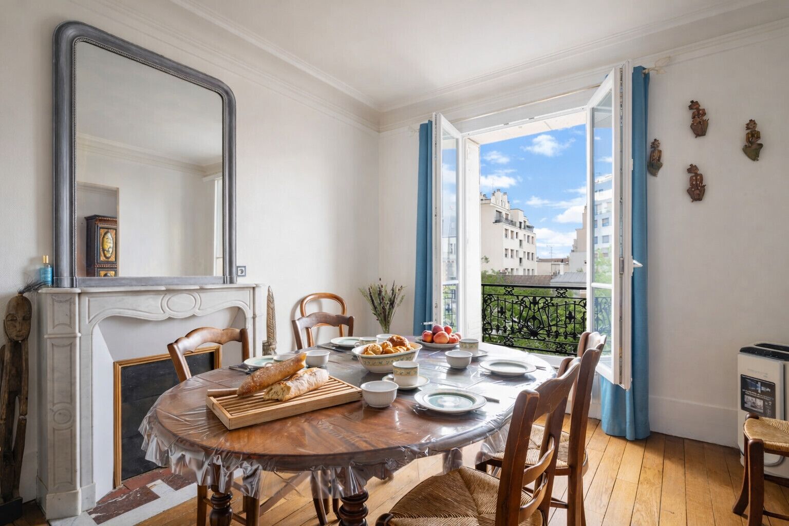Comedor con una mesa redonda, sillas, chimenea y vistas a edificios a través de ventanas abiertas.