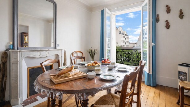 Comedor con una mesa redonda, sillas, chimenea y vistas a edificios a través de ventanas abiertas.