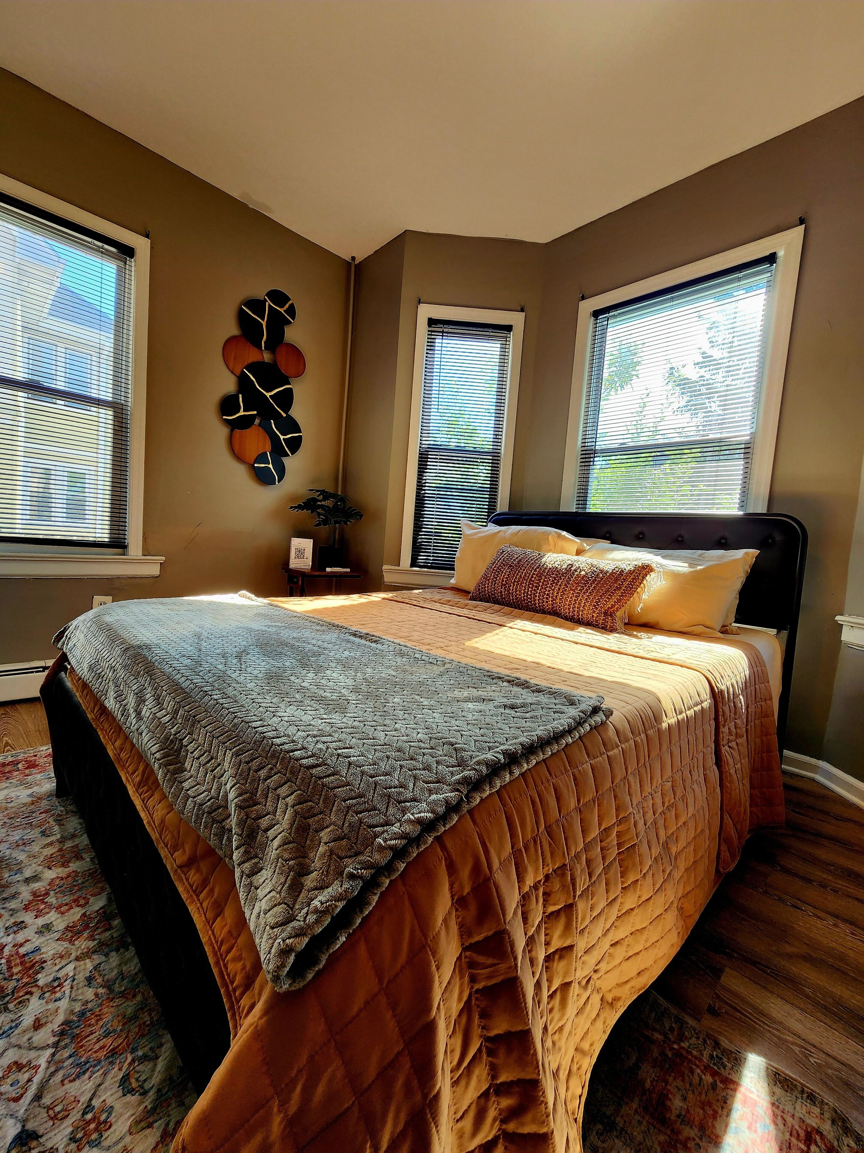 A bedroom with a large bed, two windows with blinds, a wall clock, and a decorative wall piece.