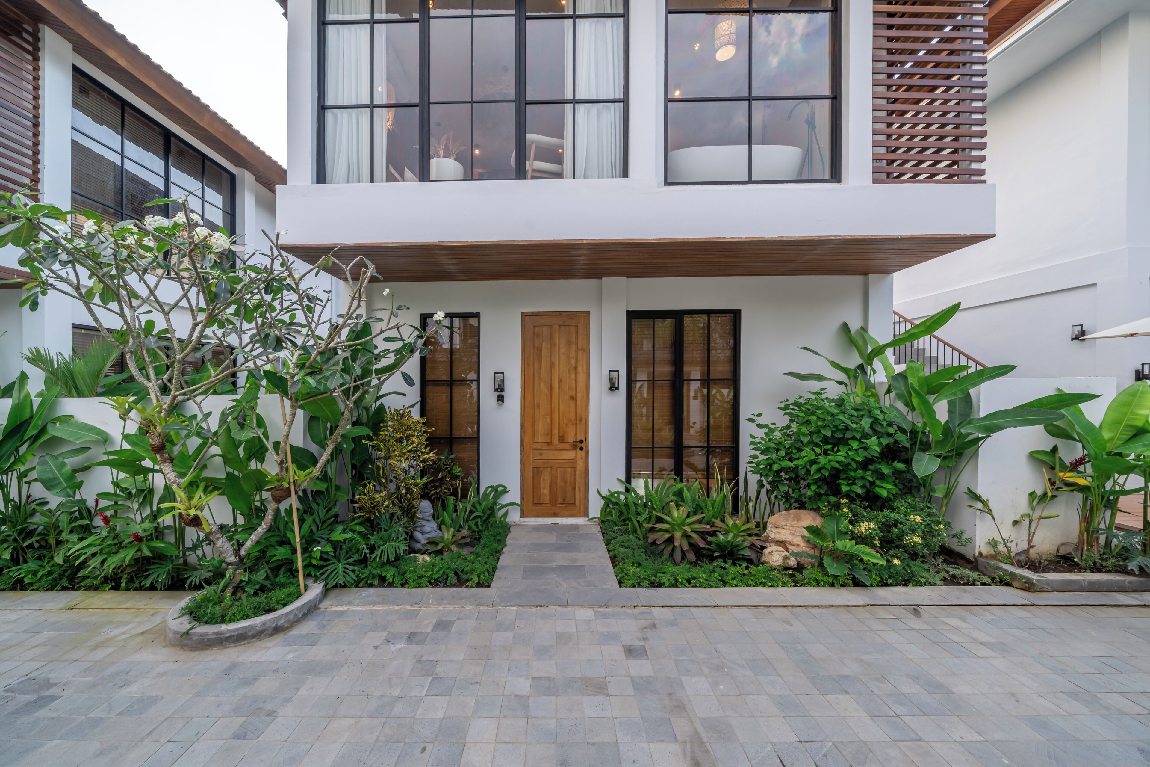 Una casa moderna de dos pisos con puerta de madera, ventanas amplias y un jardín bien cuidado con diversas plantas y un camino de piedra.