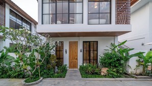 Una casa moderna de dos pisos con puerta de madera, ventanas amplias y un jardín bien cuidado con diversas plantas y un camino de piedra.