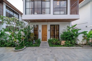 A modern two-story house with a wooden door, large windows, and a well-maintained garden with various plants and a stone pathway. - Dragonstone Villas & Suits (Gianyar)
