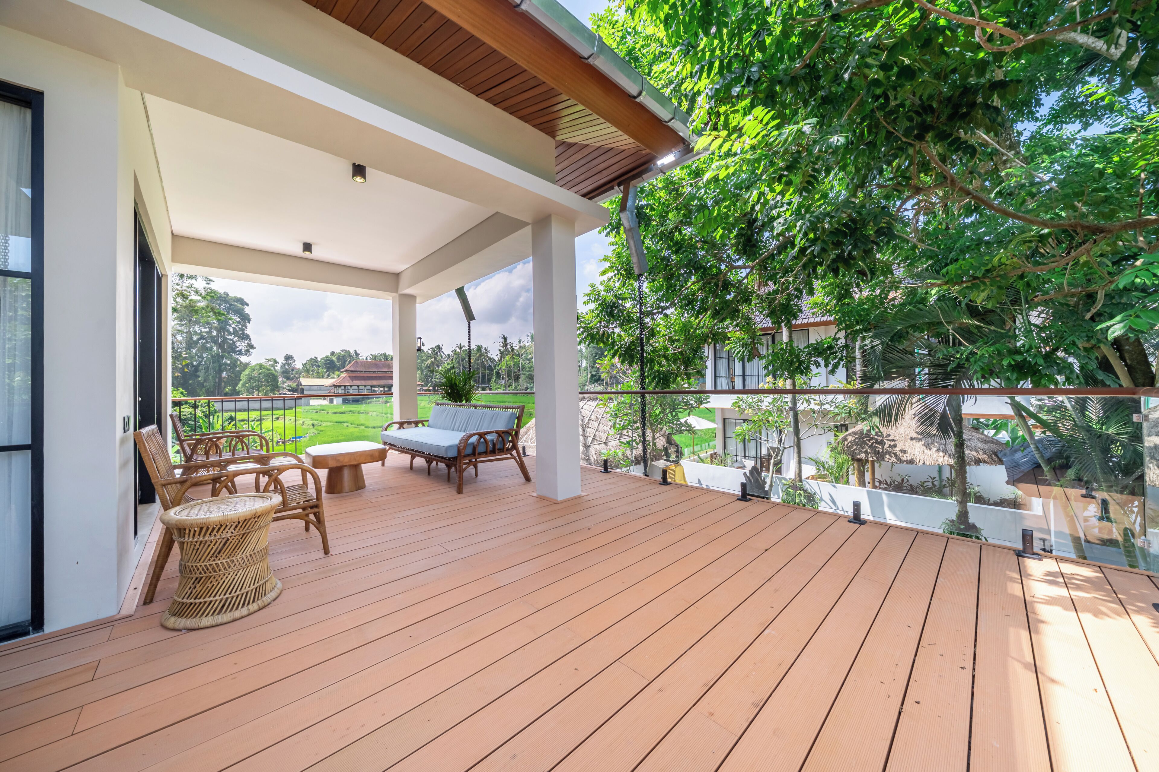 Un espacio exterior techado con terraza de madera, mobiliario de mimbre y vista a un paisaje verde.