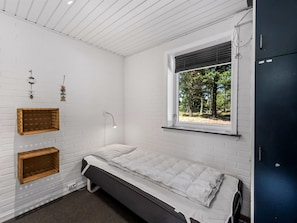 Ein Einzelbett in einem Zimmer mit weißen Backsteinwänden, einem Fenster mit Blick auf Bäume und einem dunkel gefärbten Kleiderschrank.