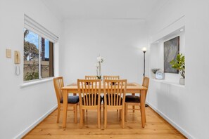 Een eethoek met een houten tafel en stoelen, een vaas met bloemen, een klok en een raam met uitzicht op bomen.