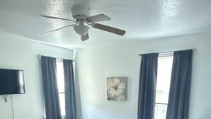 Ein Schlafzimmer mit einem Bett, einem Deckenventilator, einem Flachbildfernseher und zwei mit Vorhängen versehenen Fenstern.