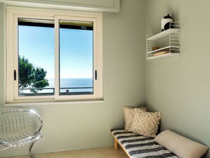 Una habitación con un ventanal grande con vistas al mar, una silla de metal, un banco con cojines y un estante blanco.