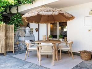 Een terras buiten met een ronde houten tafel, vier rieten stoelen, een grote rieten parasol en een stenen muur begroeid met groen.