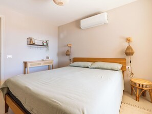 Een slaapkamer met een bed, nachtkastje, wandairconditioner en een klein rond krukje.