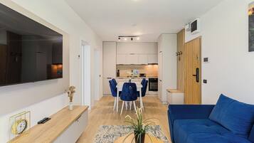 A modern living room with a blue sofa, a dining area with chairs, and a kitchen in the background.