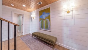 A hallway with a wooden ceiling, a bench, a patterned rug, and a painting on the wall.