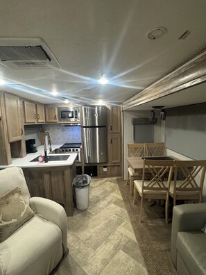 Une cuisine compacte intégrée au camping-car, comprenant un réfrigérateur, un four à micro-ondes, un évier et une cuisinière. Il y a également un coin repas avec une table et des chaises.